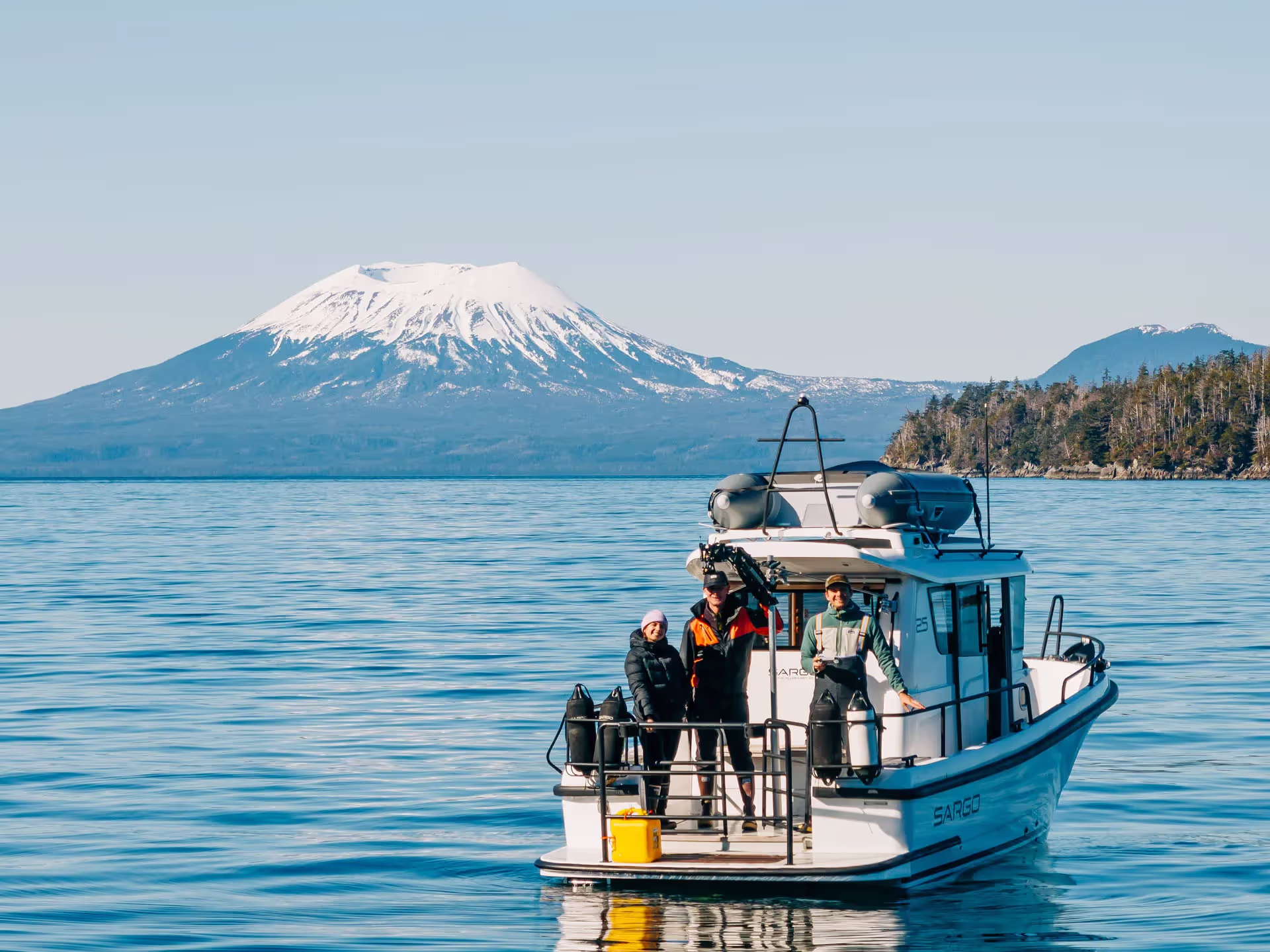 Three people standing on the deck of a white boat on calm water with a snow-capped mountain and forested hills in the background.