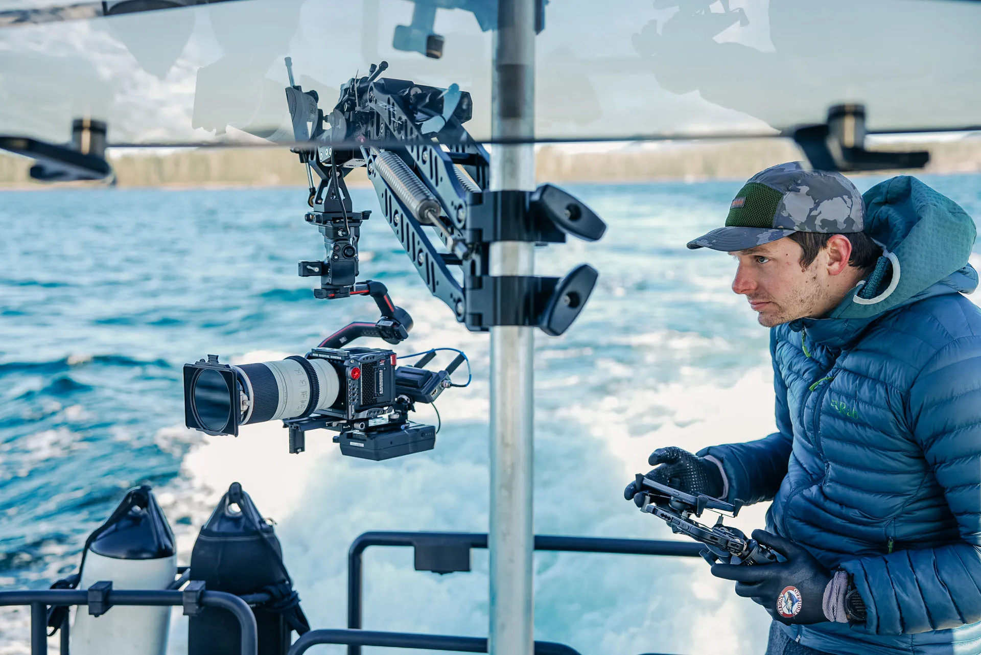 Man wearing a blue jacket and camouflage cap operating a camera mounted on a boat overlooking the water.