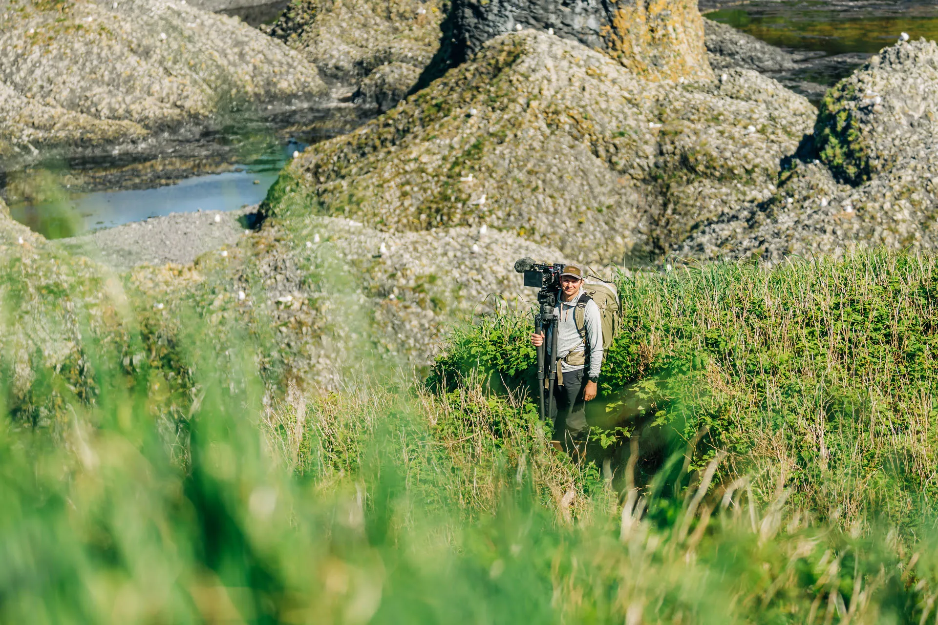 Ben Hamilton filmmaker wearing backpack and cap holding a professional video camera on tripod standing in green grass with rocky terrain in the background.