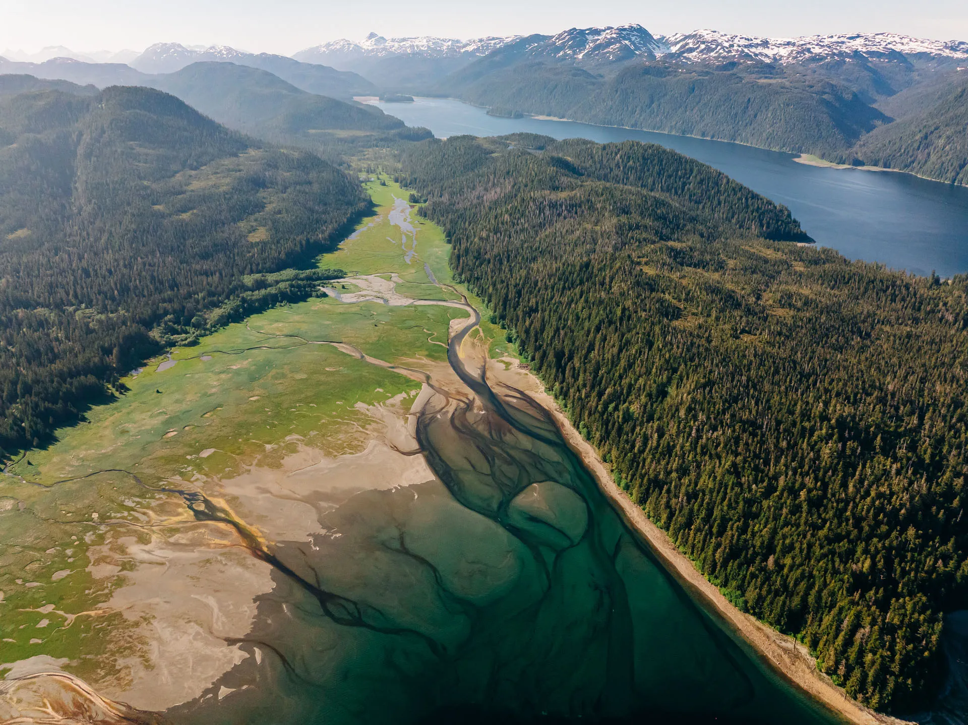 Aerial view of a river delta flowing into a lake, surrounded by dense forested mountains with snow-capped peaks in the distance.