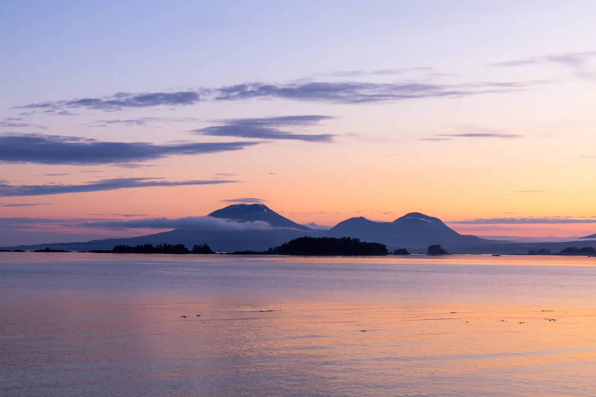 Calm body of water reflecting pastel colors of sunrise with silhouetted islands and distant mountains in Sitka Sound.