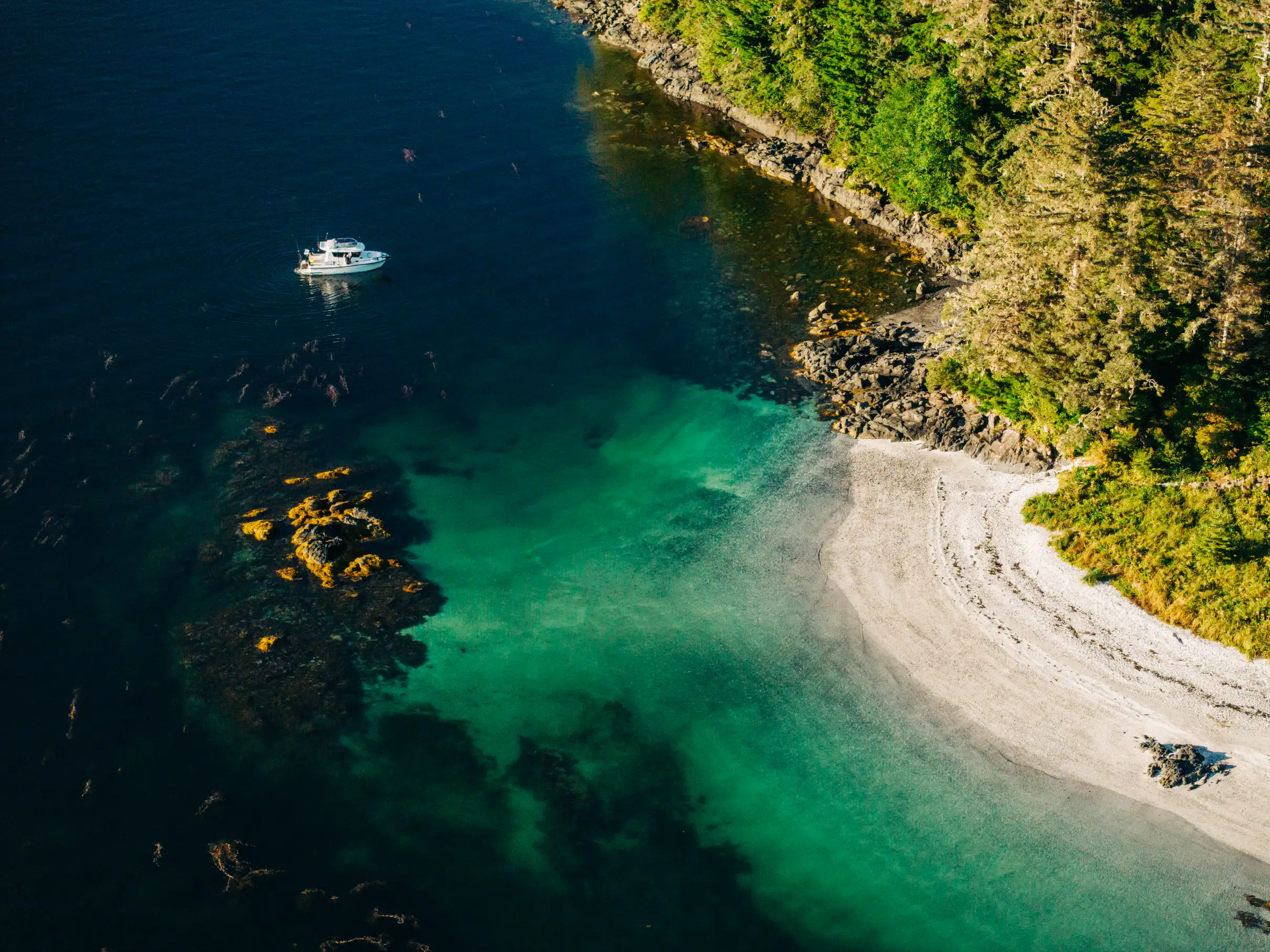 Aerial view of a small white boat floating near a rocky shoreline with clear turquoise water and dense green forest in Sitka Sound.