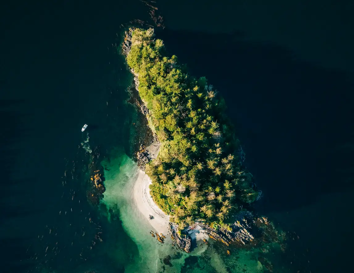 Aerial view of a small, densely forested island with clear turquoise water and a white sandy beach, surrounded by dark blue ocean in Sitka Sound.