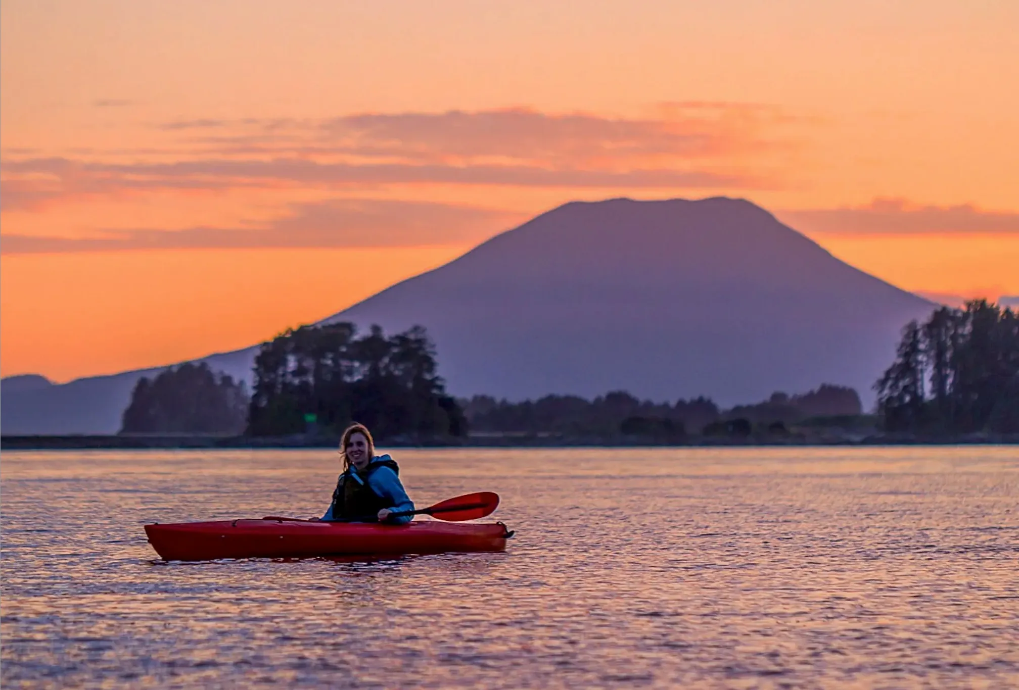 Person kayaking on calm water at sunset with Mt. Edgecumbe the volcano behind them in the distance.