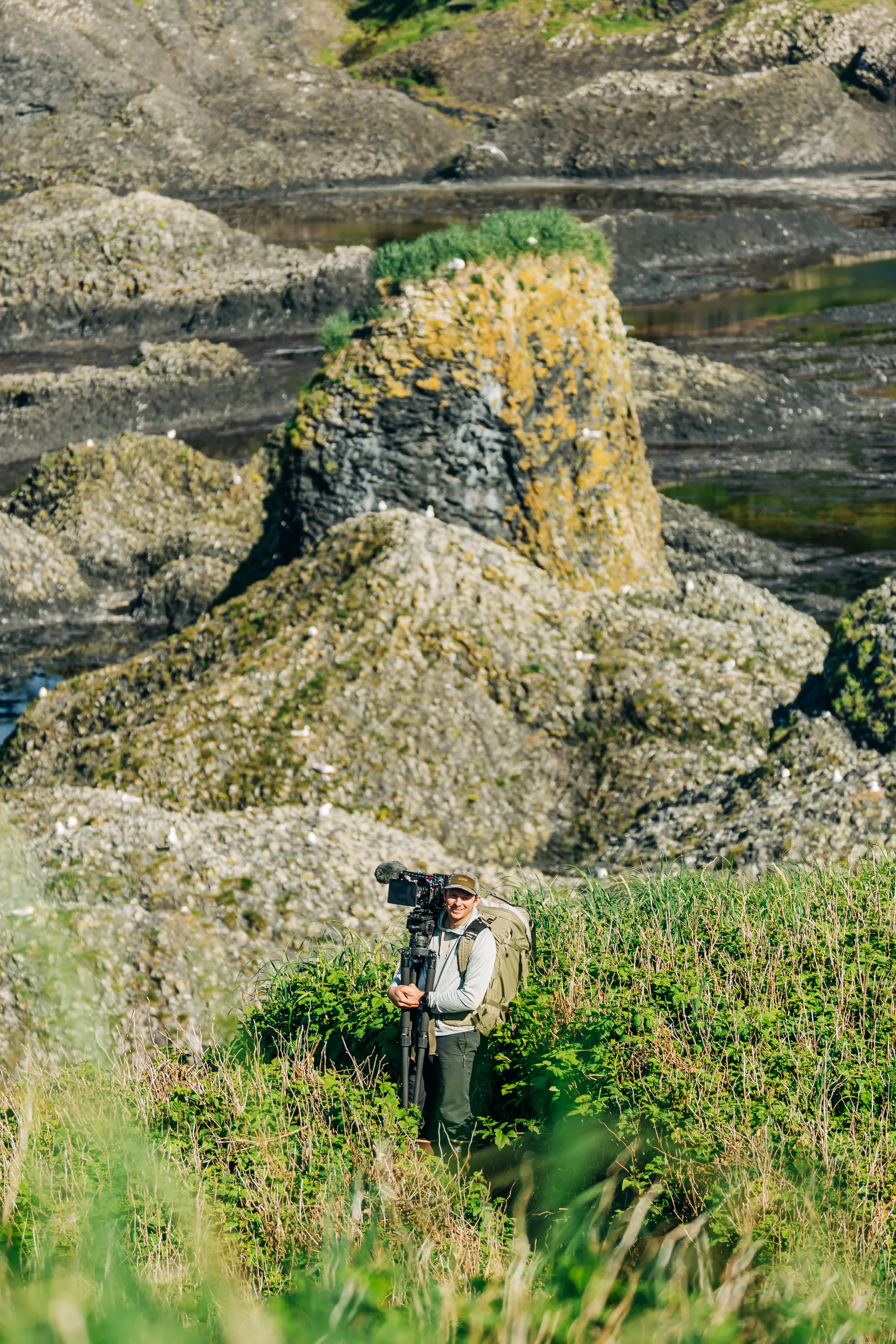 Photographer standing among green bushes near rocky coastline holding a professional camera on a tripod.