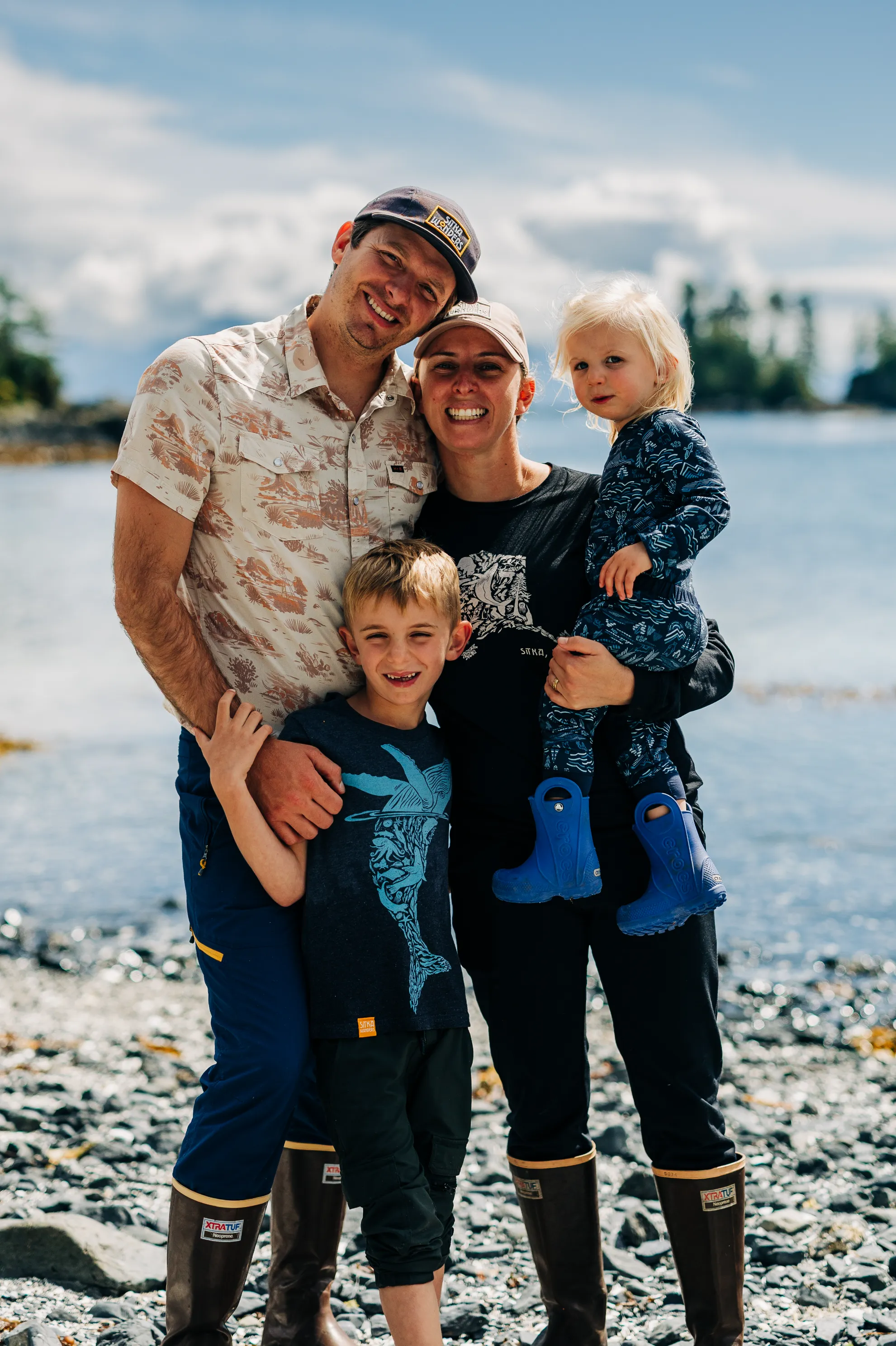Smiling family of four standing on a rocky shoreline with water and trees in the background, wearing outdoor clothing and boots.