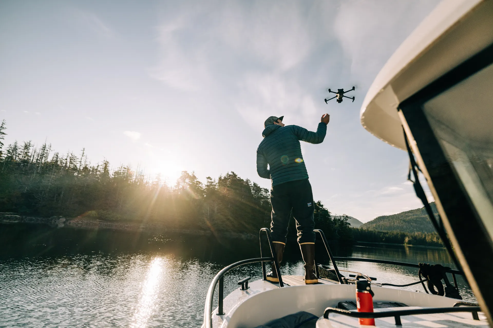 Person standing on a boat launching a drone over calm water with forest and mountains in the background at sunset.