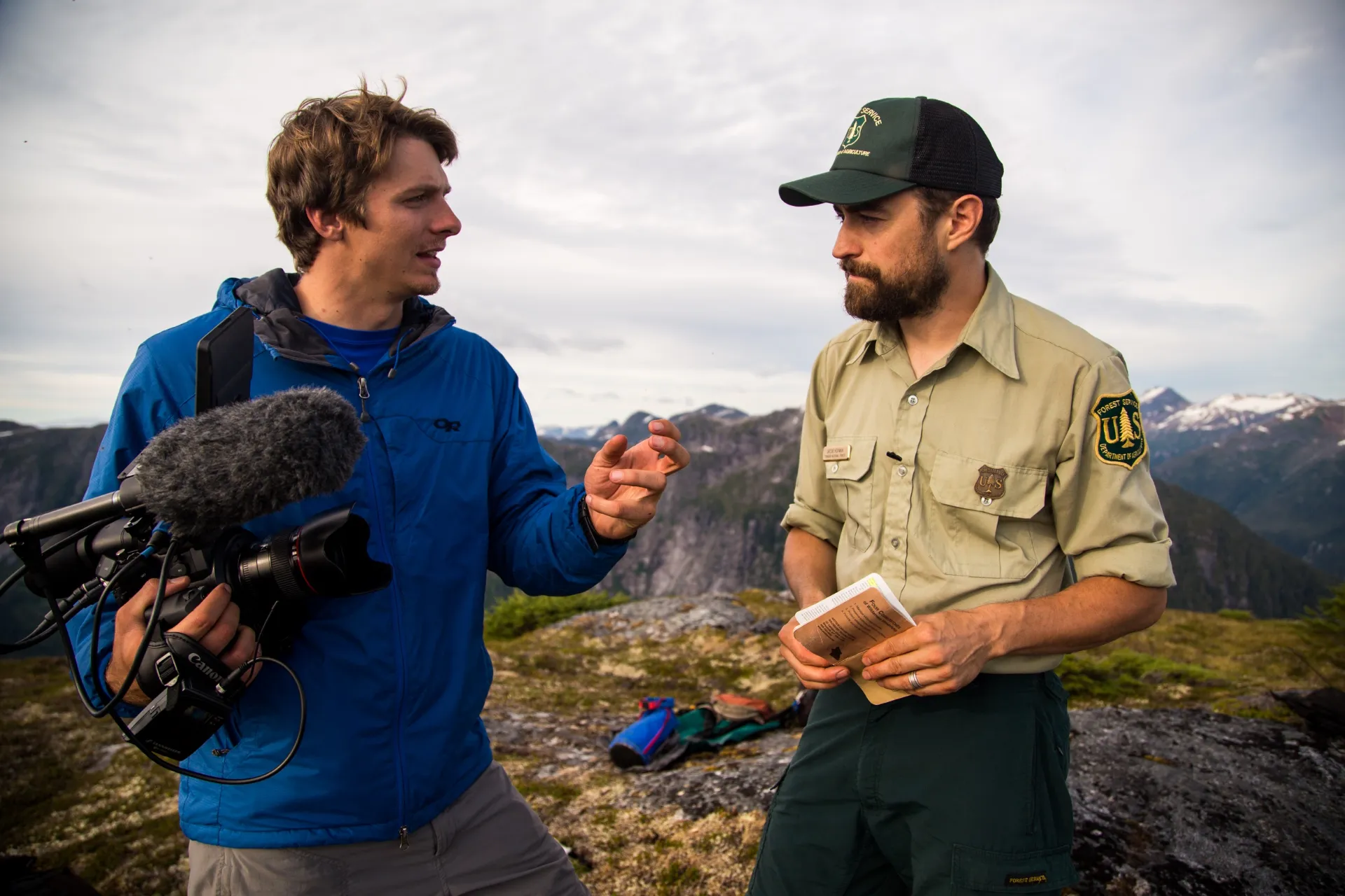 A man in a blue jacket holding a video camera interviews a man in a US Forest Service uniform outdoors with mountains in the background.