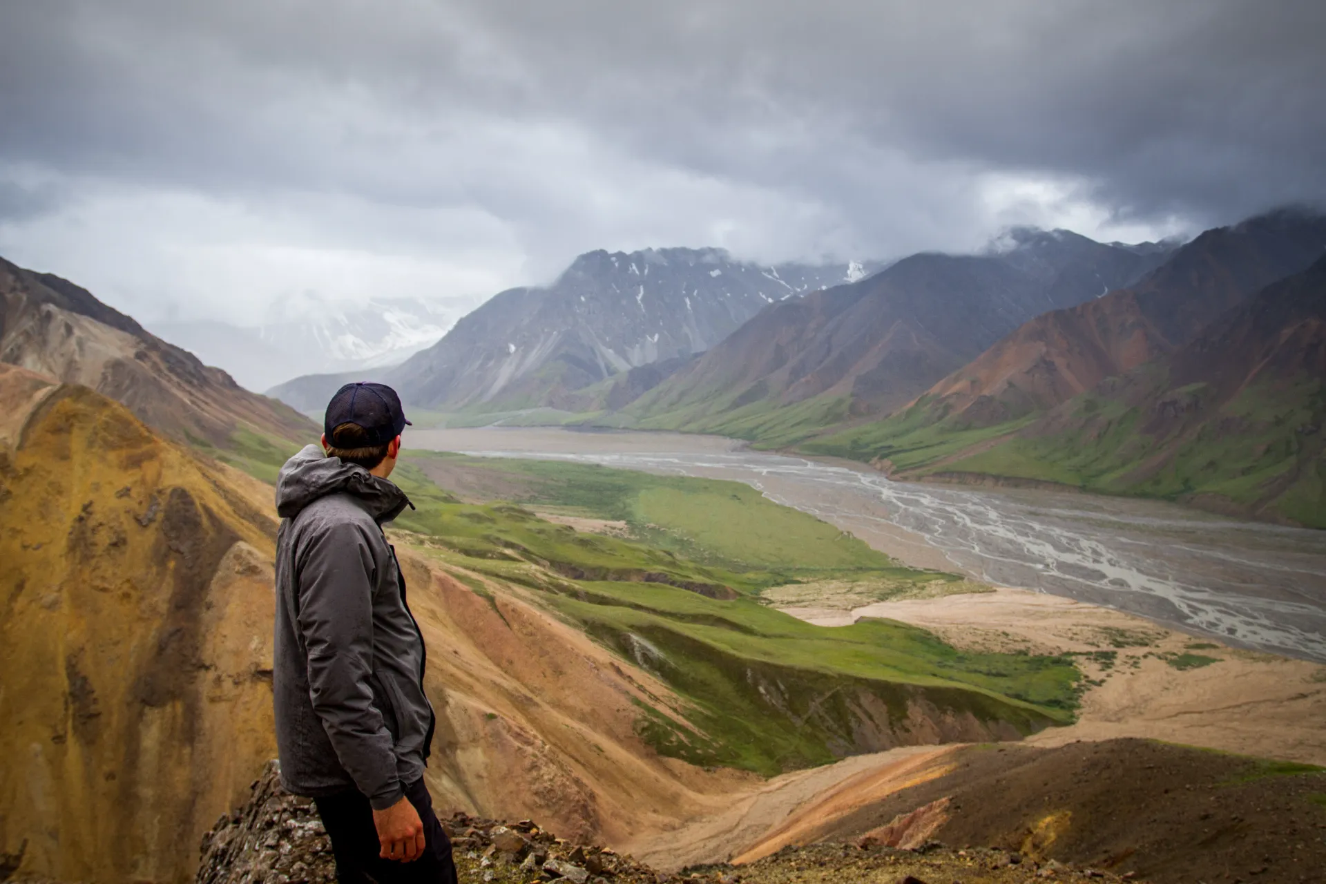Person in a jacket and cap overlooking a river valley surrounded by rugged mountains under cloudy skies.