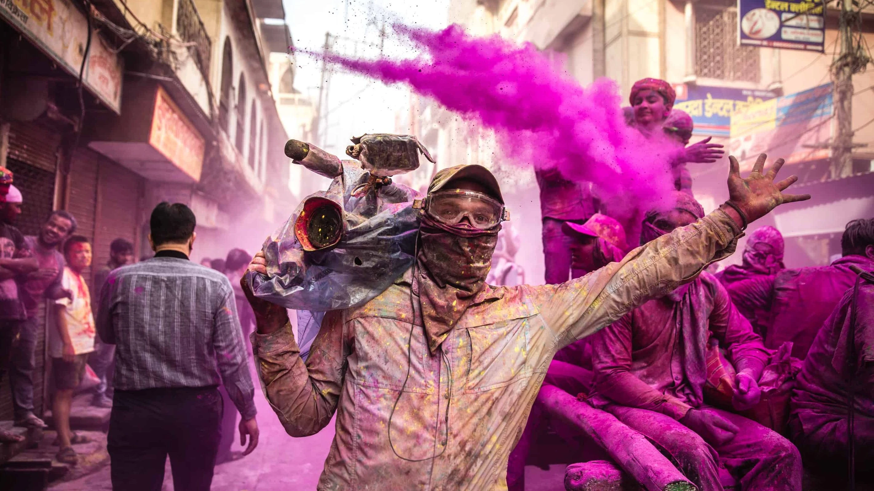 Person wearing goggles and face covering spraying vibrant pink powder during a crowded street Holi festival celebration.