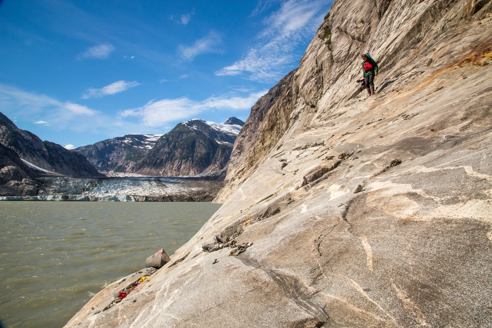 Person climbing steep rocky slope overlooking a lake with a glacier and snow-capped mountains in the background under a blue sky.