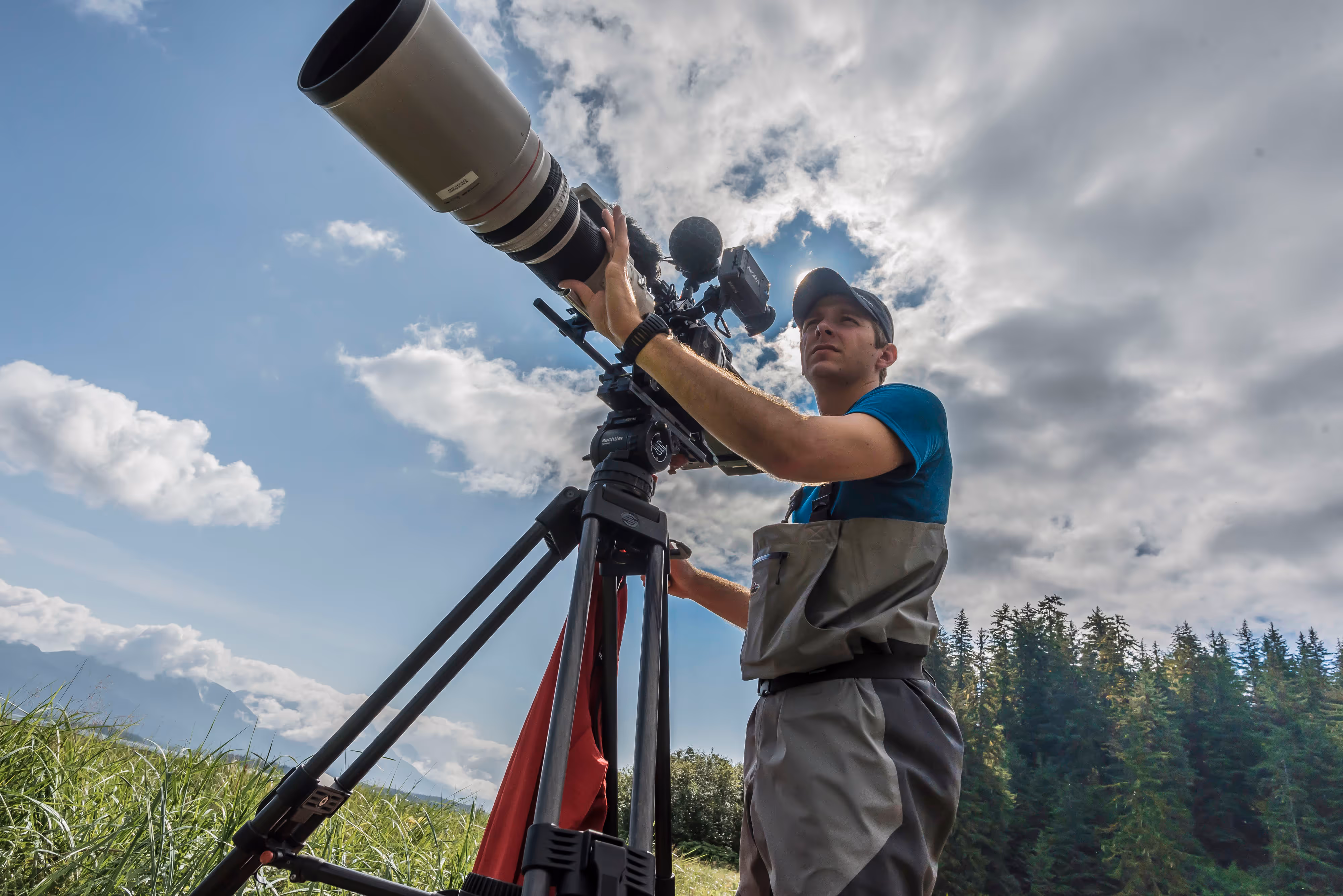 Man wearing outdoor gear filming with a large telephoto camera lens on a tripod in a grassy field with forest and cloudy sky background.