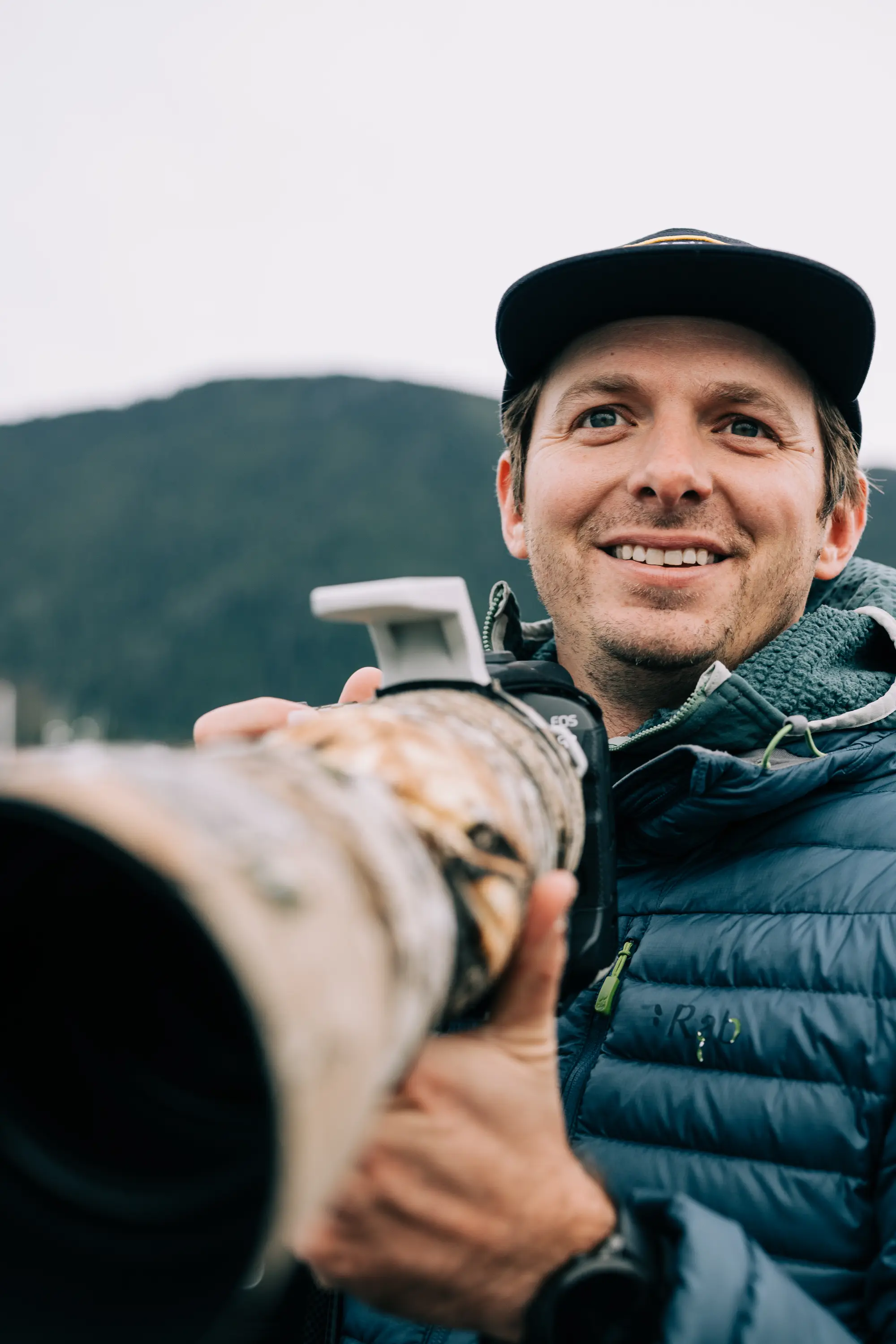 Ben Hamilton smiling and wearing a cap and blue jacket holding a camera with a large camouflage telephoto lens outdoors.