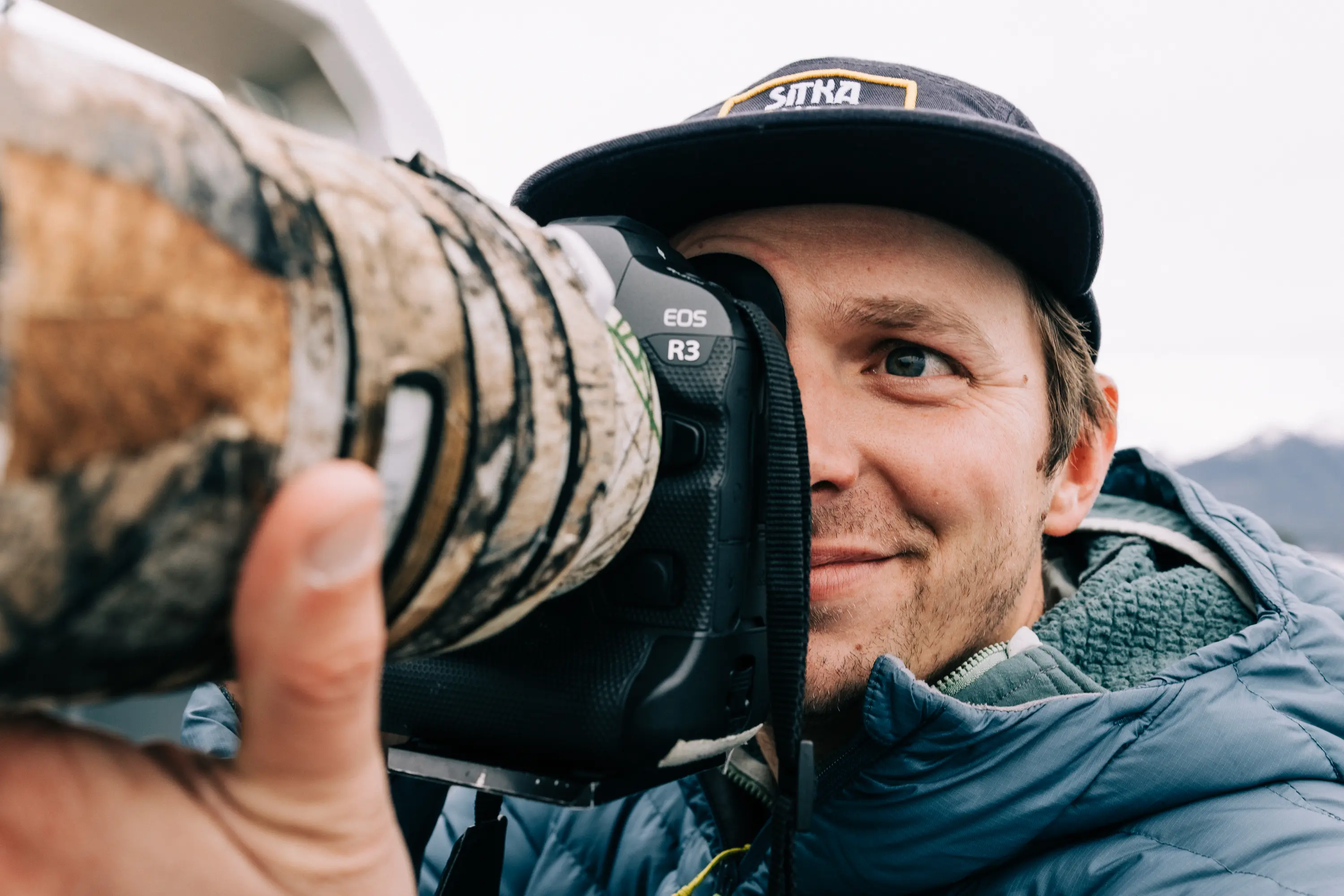 Man in a blue jacket and black Sitka cap holding a Canon EOS R3 camera with a camouflage lens cover, looking through the viewfinder.