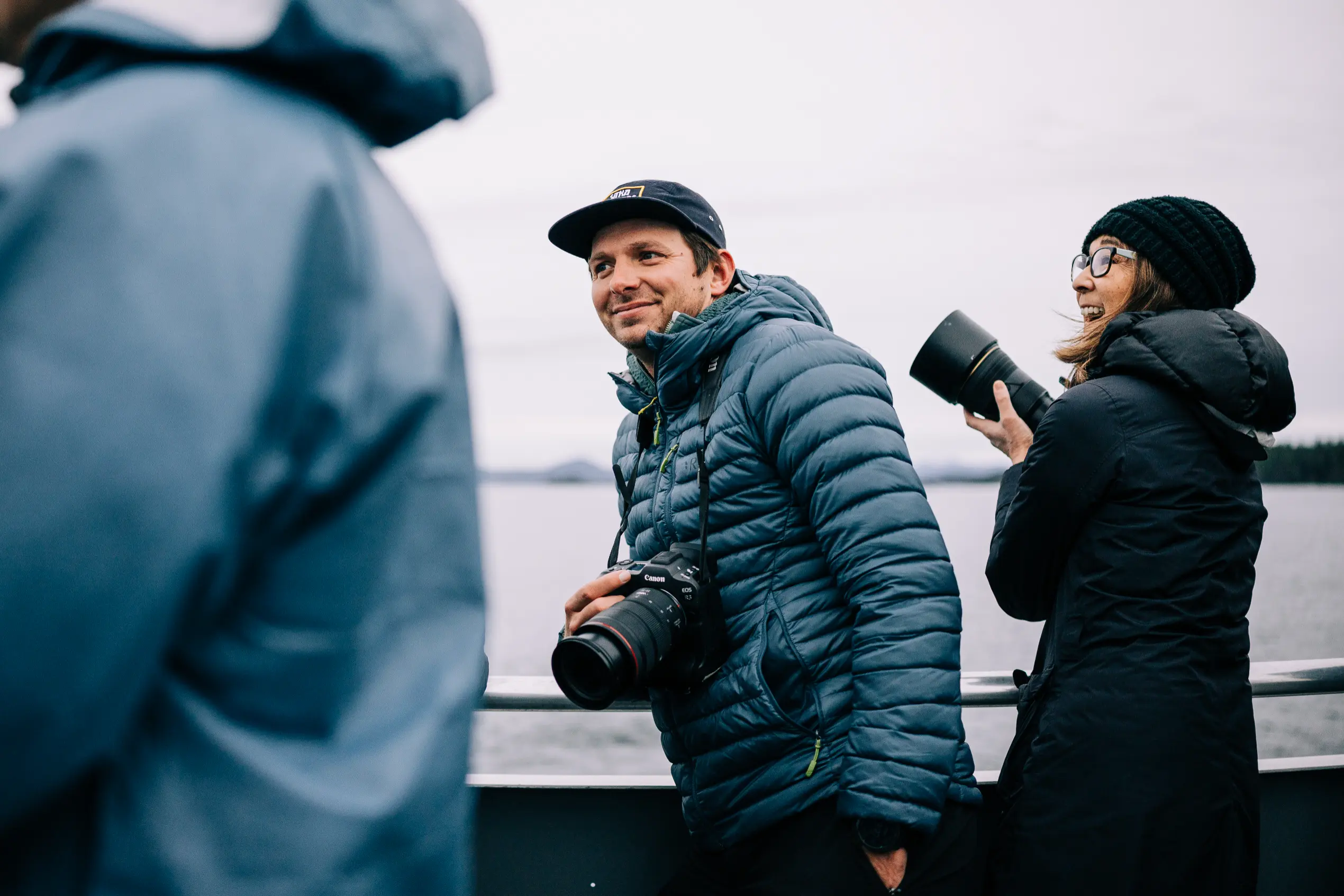 Two photographers, a man and a woman, wearing jackets and holding cameras on a boat with water and cloudy sky in the background.