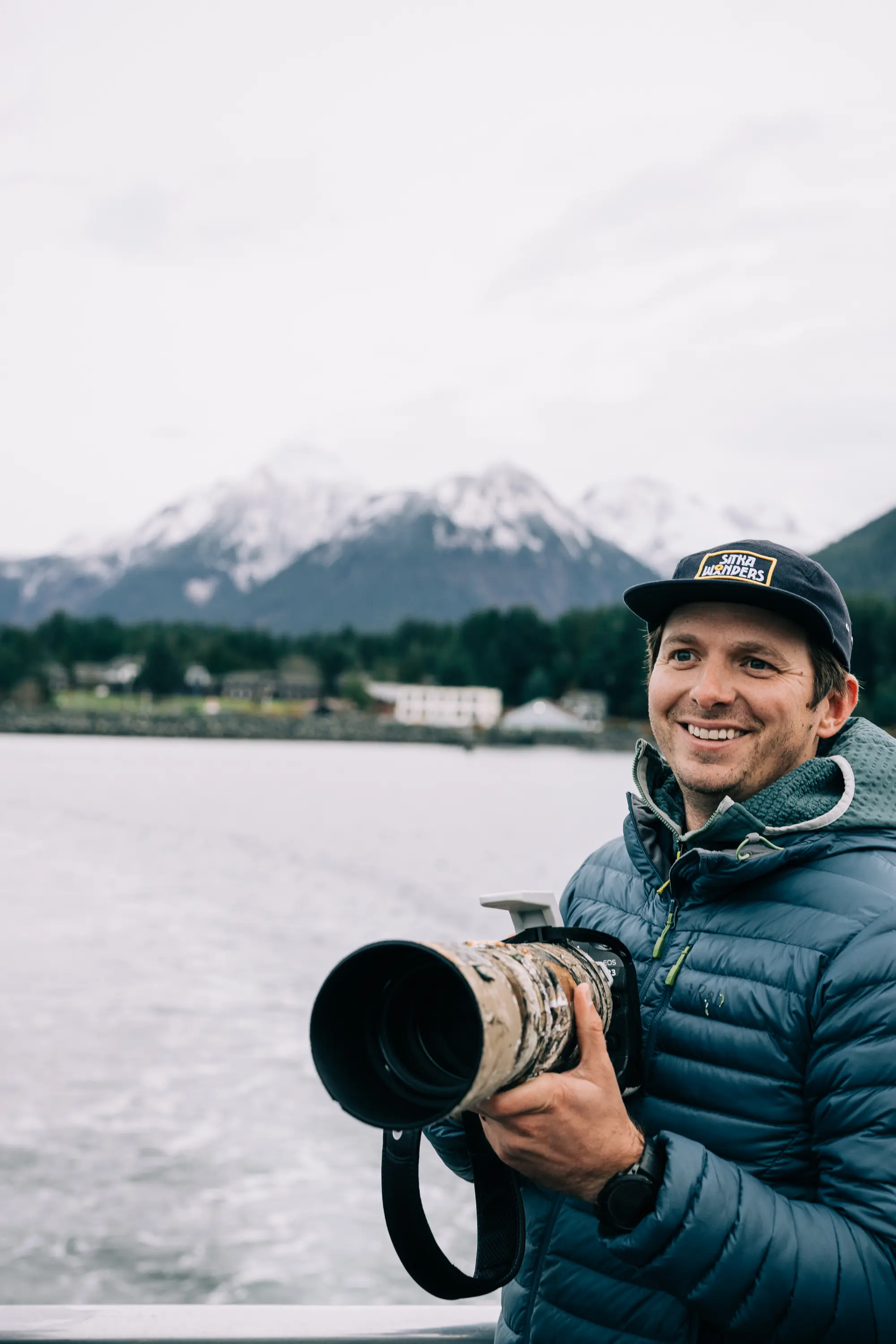 Smiling man in a blue jacket holding a professional camera with a large lens near a lake with snow-capped mountains in the background.