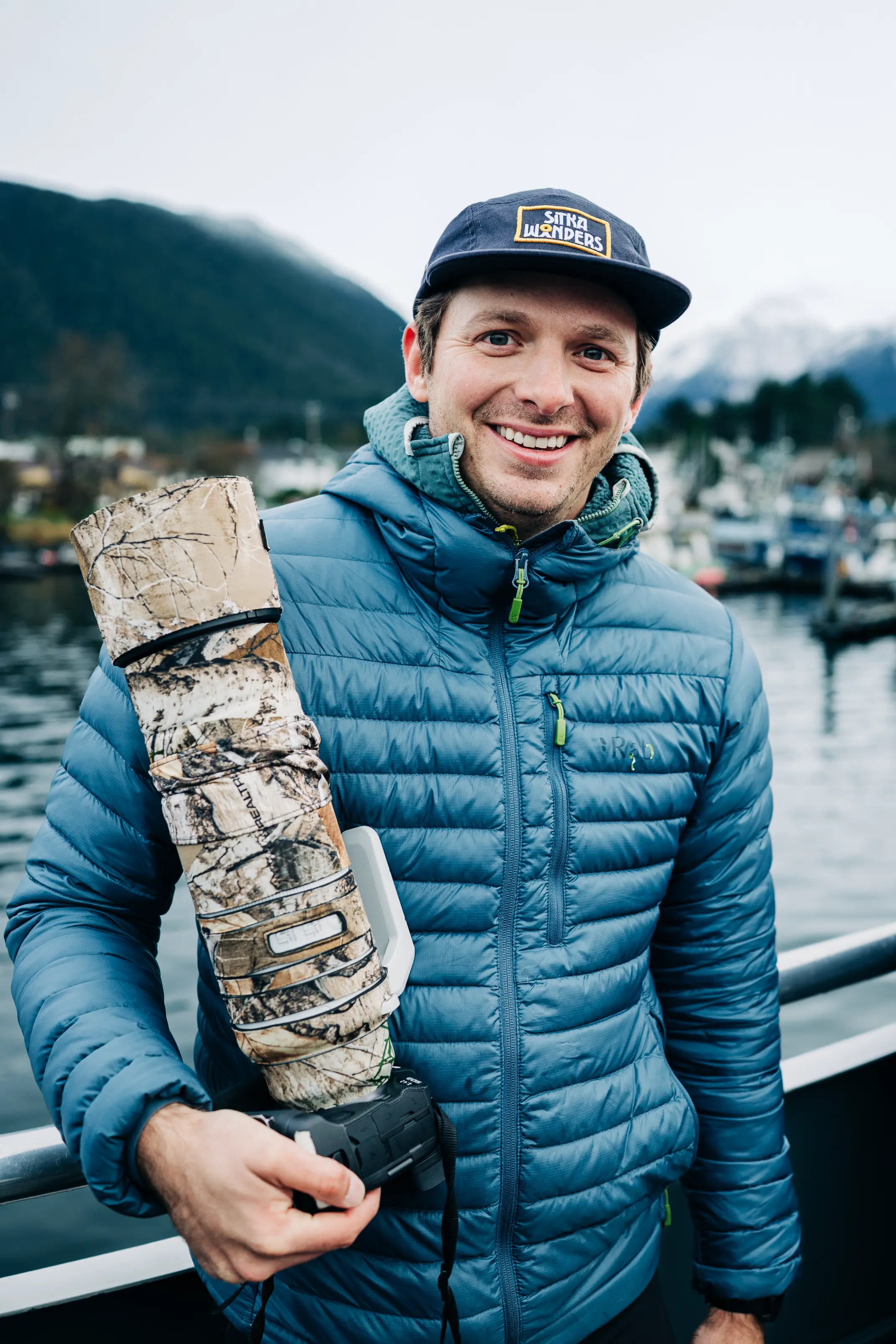 Smiling man in a blue puffer jacket and navy Sitka Wonders cap holding a camera with a camouflage lens cover, standing outdoors near water and mountains.