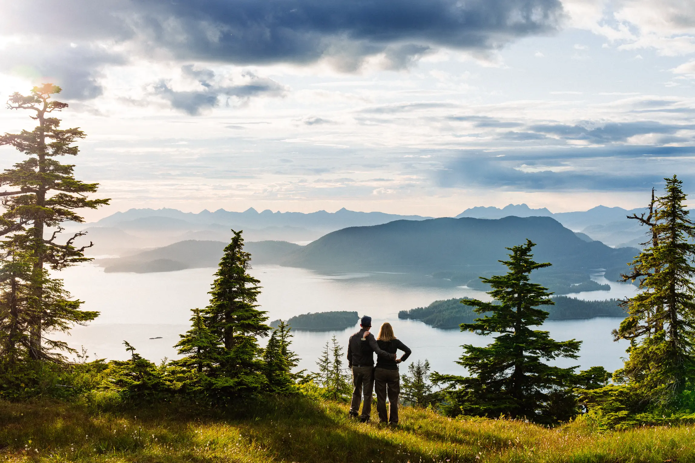 A couple stands on a mountain ridge above Sitka Sound at sunset, looking out over islands and distant peaks