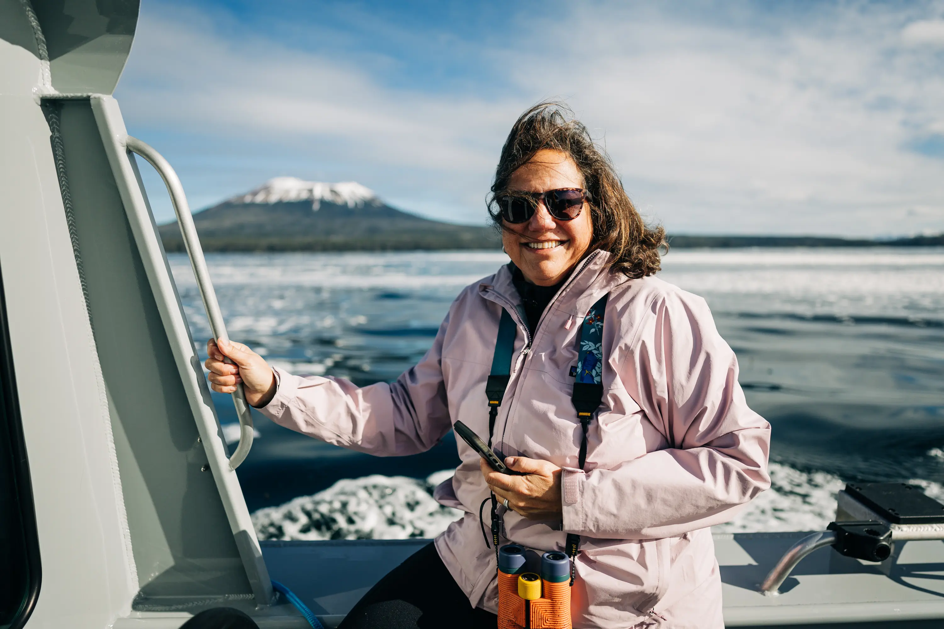 A smiling guest on the boat with binoculars, Mount Edgecumbe in the background