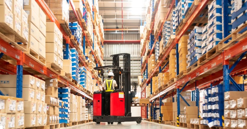 a warehouse worker riding a forklift passes a bunch of boxes filed