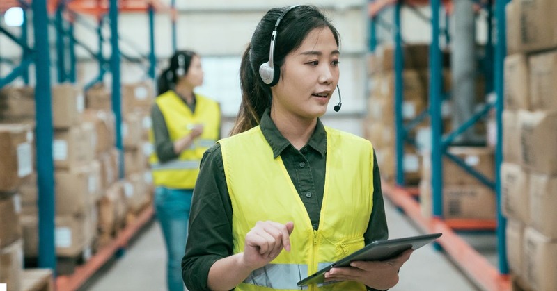 a woman in a warehouse talking on the headset while holding a tablet