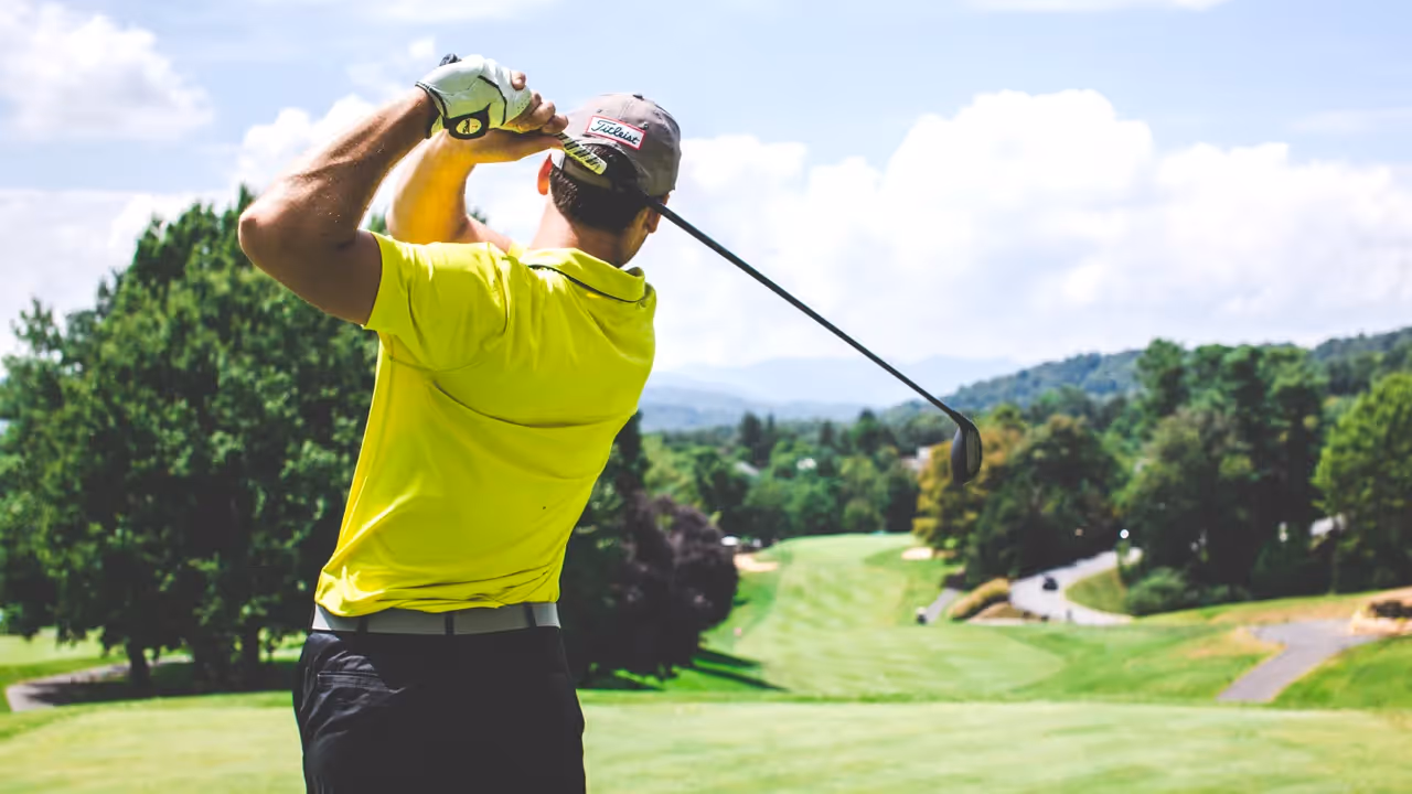 Man in a yellow shirt mid golf swing at a golf course.