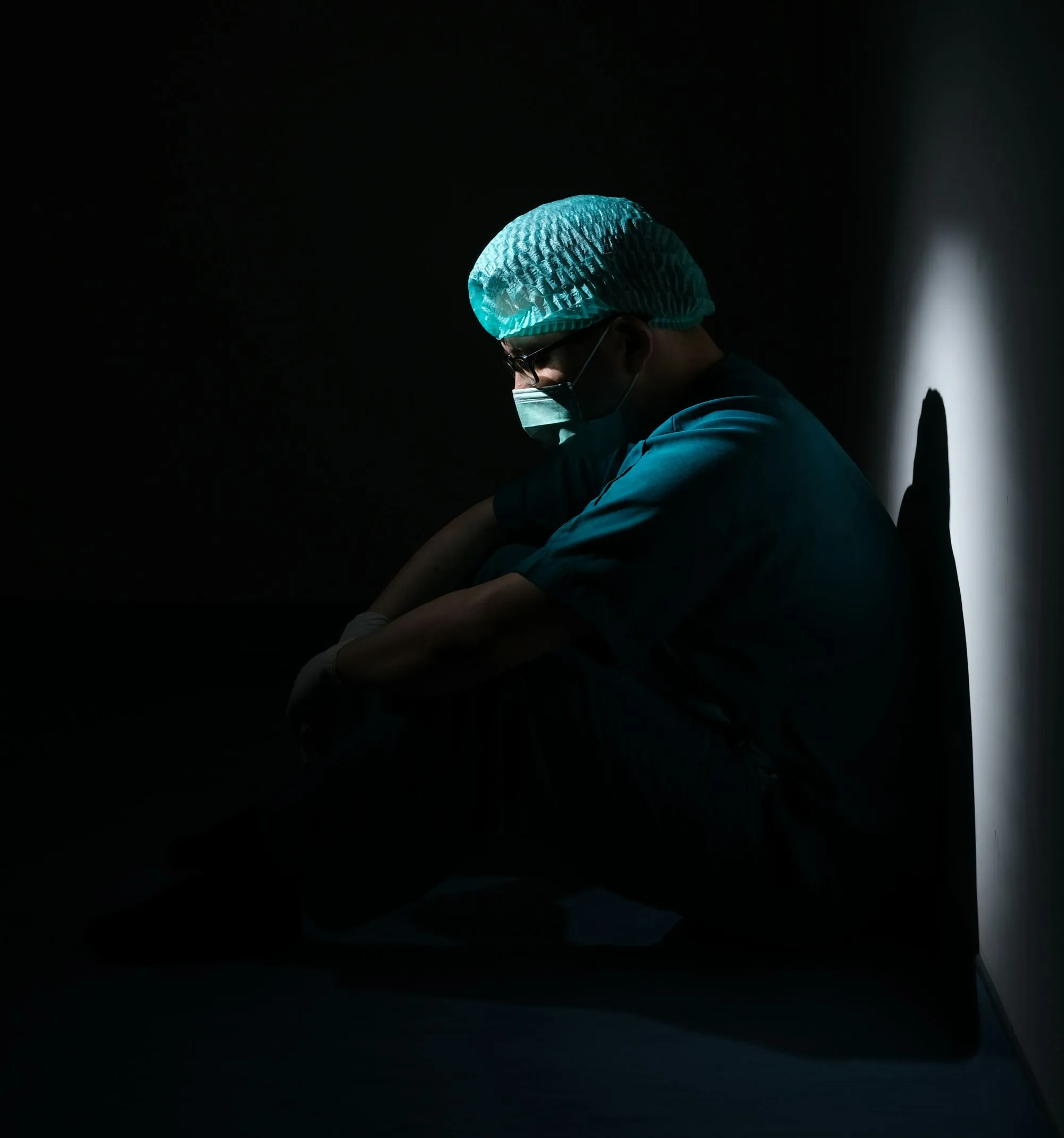 A physician is sitting with his back to the wall in the dark looking exhausted.