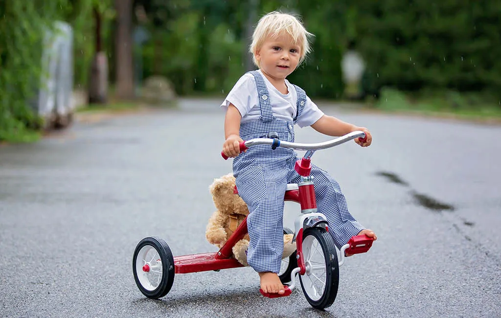 Cute little boy barefoot, riding a tricycle in the rain with a teddy bear.