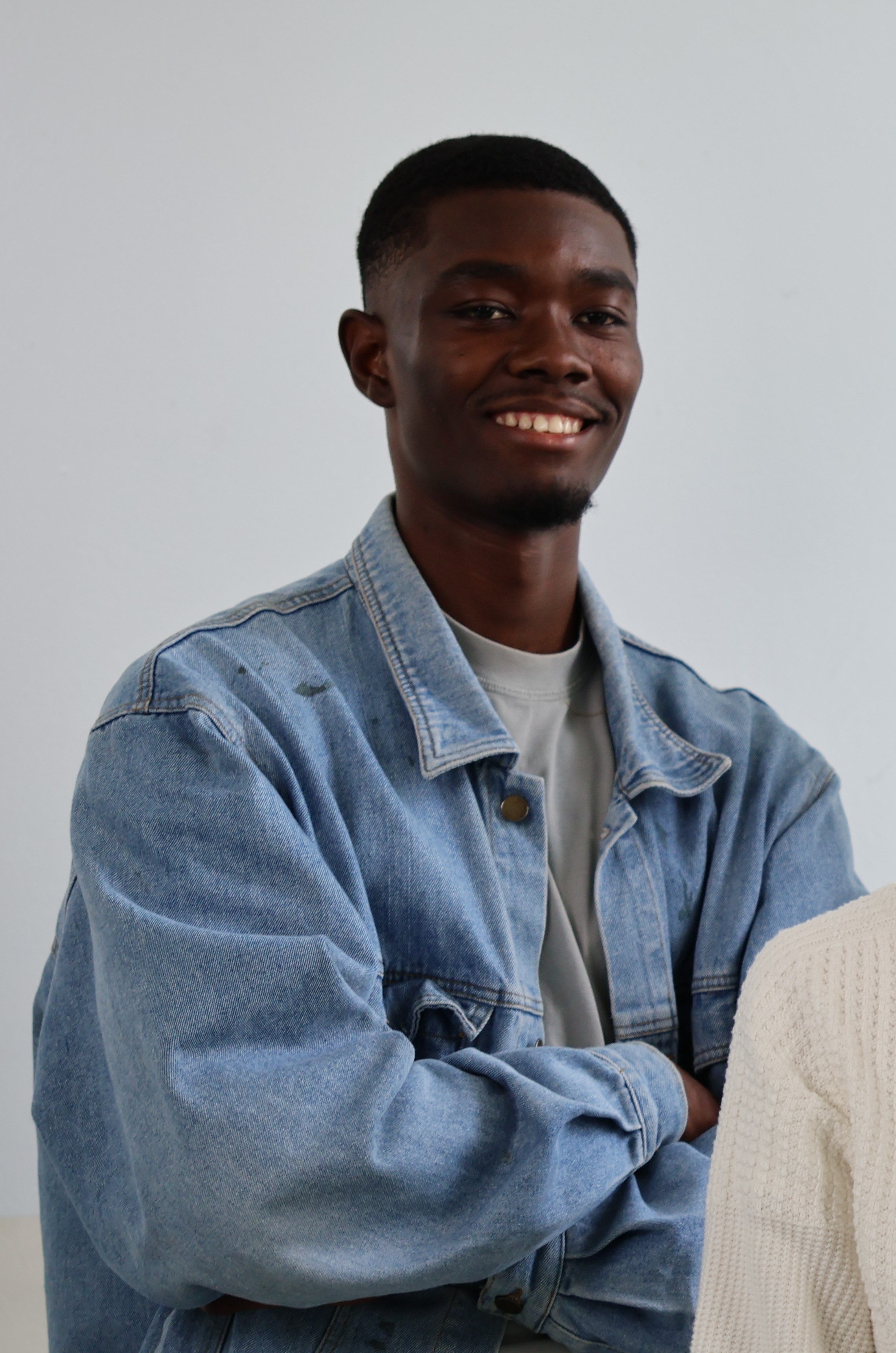 Smiling man wearing a light blue denim jacket with arms crossed against a plain light gray background.
