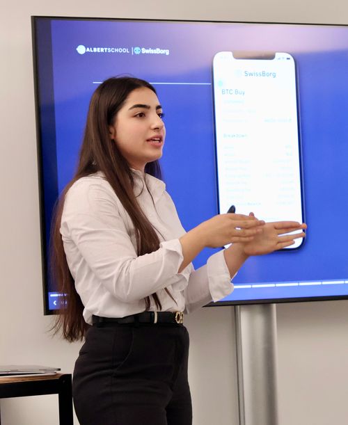 A young woman with long dark hair in a white shirt and black pants is giving a presentation in front of a screen displaying a SwissBorg app interface.