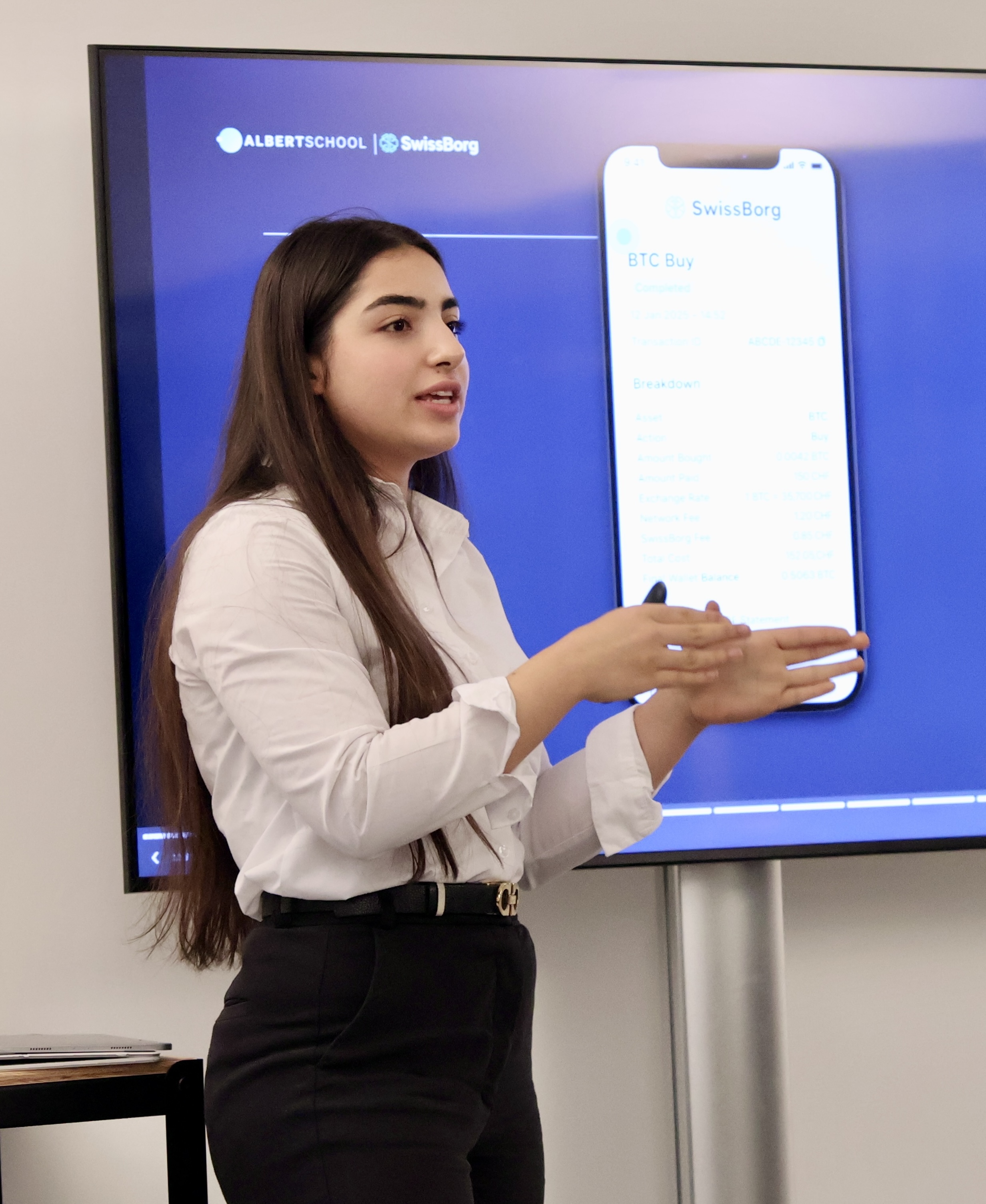 A young woman with long dark hair in a white shirt and black pants is giving a presentation in front of a screen displaying a SwissBorg app interface.