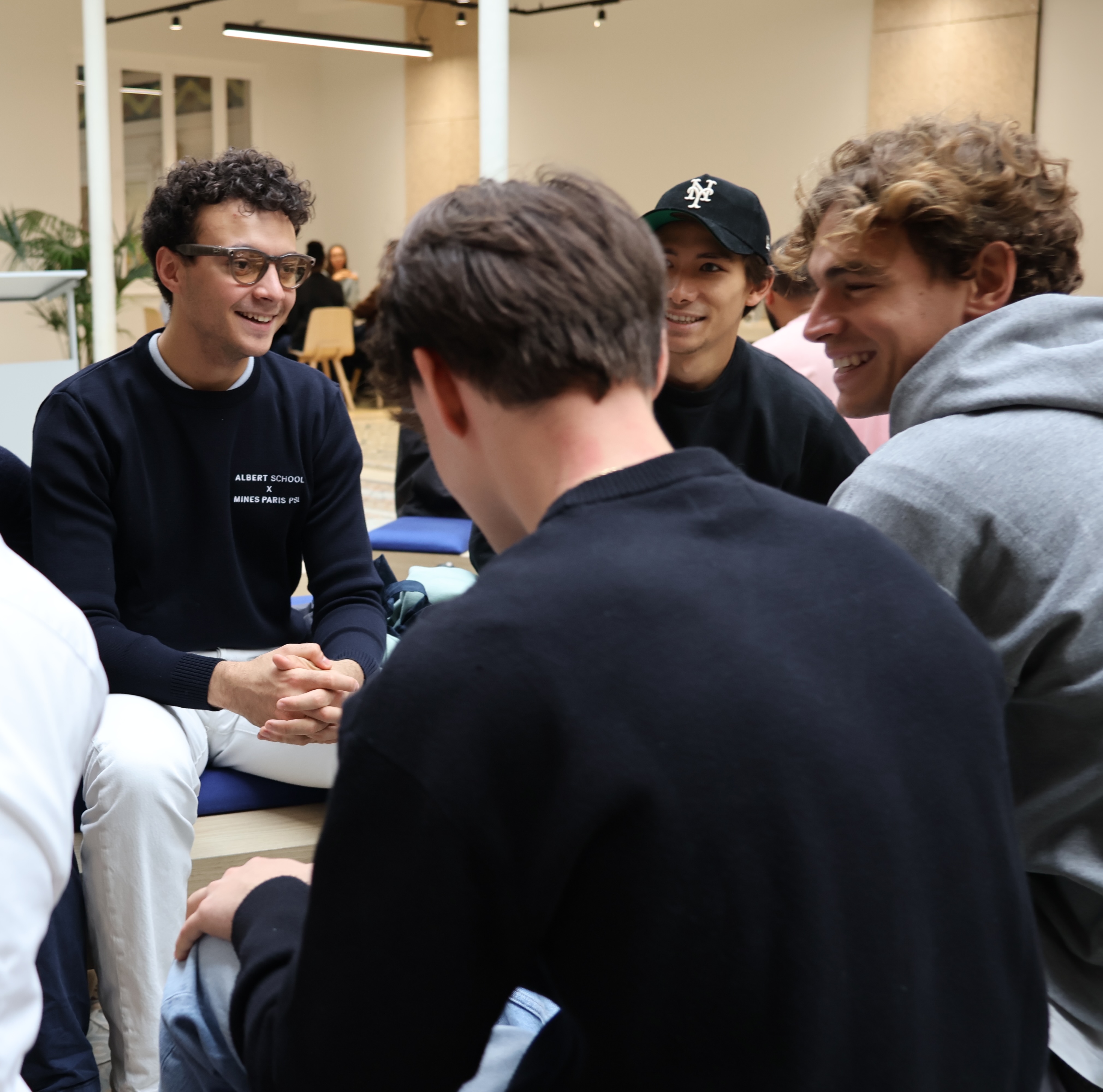 Group of young men sitting and chatting indoors, with one wearing glasses and a navy sweater that reads Albert School x Mines Paris.