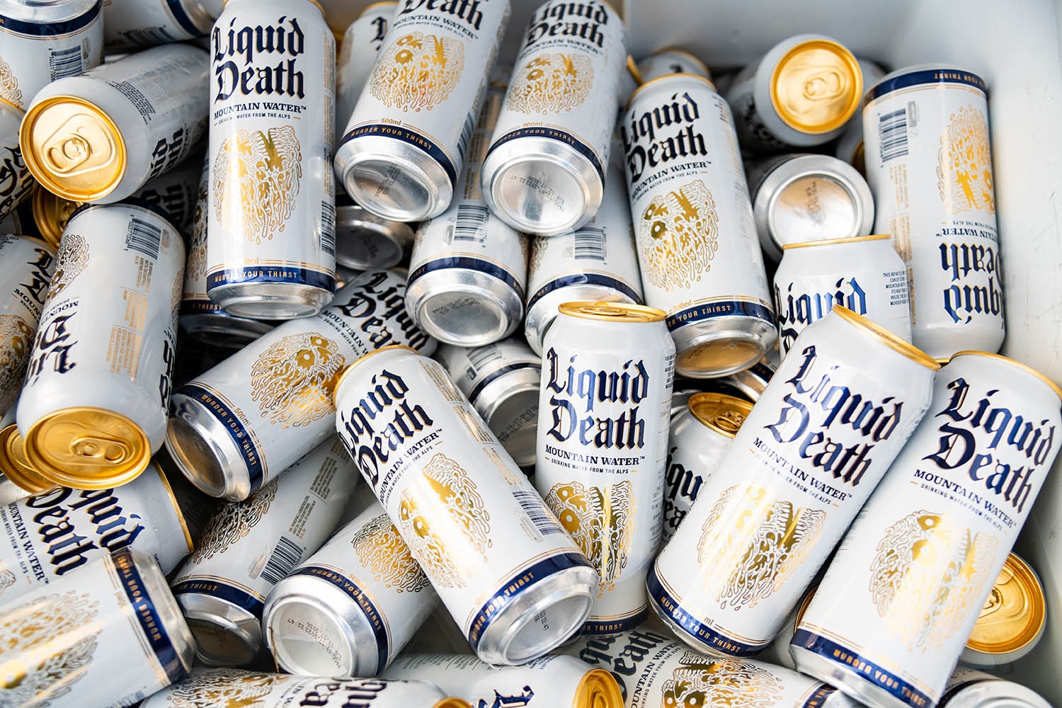 Pile of Liquid Death Mountain Water cans showing the brand's distinctive white-and-gold gothic packaging. The bold, rebellious visual identity is what makes Liquid Death one of the most mentally available beverage brands in its category.