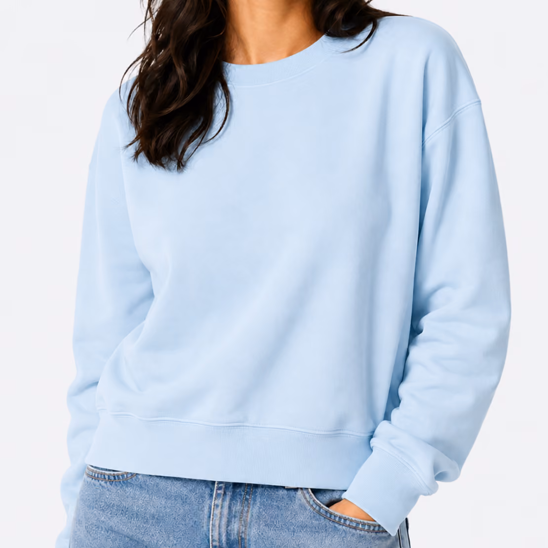 Woman wearing a light blue crewneck sweatshirt with long sleeves and ribbed cuffs, paired with blue denim jeans, with one hand in her pocket.