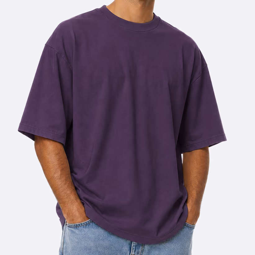 Man wearing a loose-fitting, solid dark purple short-sleeve t-shirt with hands in light blue jeans pockets against a plain light background.