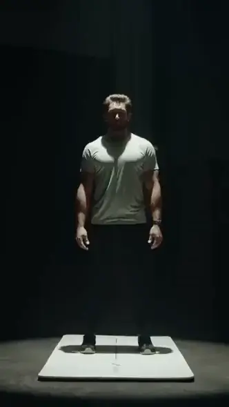 "Man standing alone on a lit platform in a dark room, wearing a light-colored t-shirt and dark pants, facing forward with arms relaxed at his sides