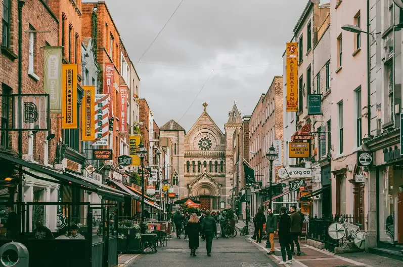 Pedestrian street lined with shops and restaurants leading to a large historic church under a cloudy sky.