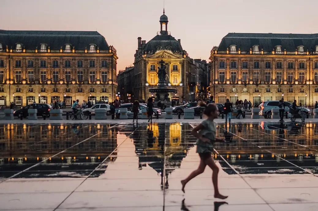 A child running barefoot across a reflective water surface with historic lit buildings and a statue in the background at dusk.