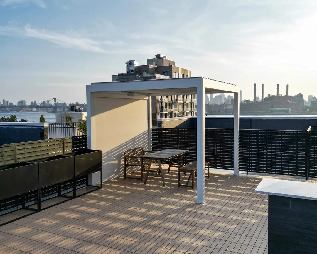 Composite tile rooftop deck with white pergola, dining area, and privacy screens overlooking the NYC skyline and East River