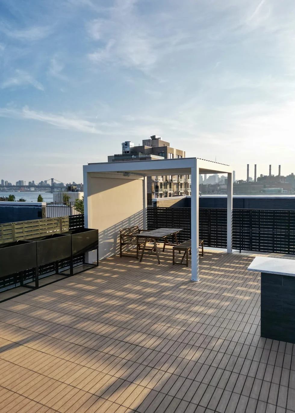 NYC rooftop deck featuring composite decking tiles, a white aluminum pergola with dining furniture, metal planters, and slatted privacy screens with waterfront skyline views