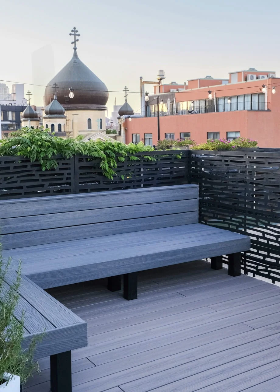 Brooklyn rooftop deck featuring gray composite decking, L-shaped built-in planters with mixed greenery, black decorative metal privacy panels, and string lights
