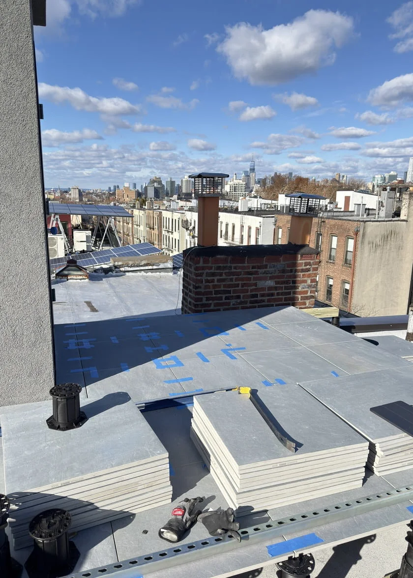 Porcelain pavers being installed on a pedestal system on a Brooklyn rooftop, with stacked tiles, blue waterproofing membrane, and city skyline views