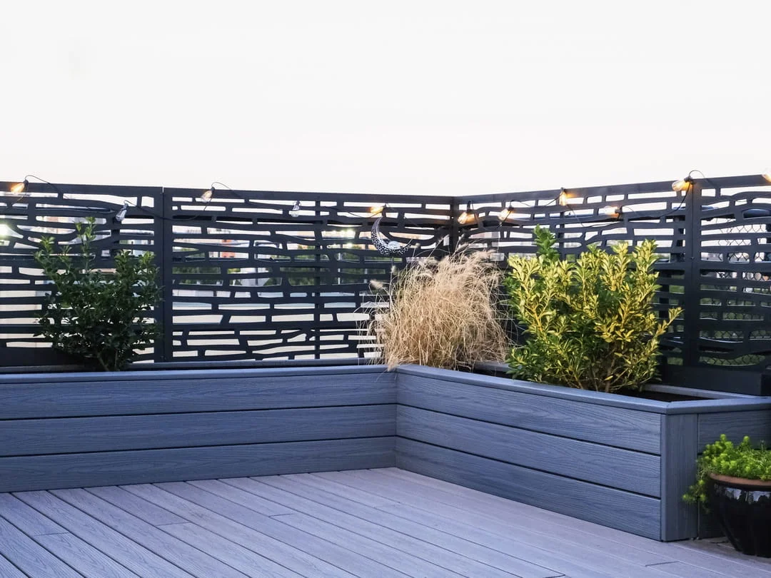 Gray composite built-in planter boxes with mixed shrubs and ornamental grasses behind decorative black metal privacy screens with string lights