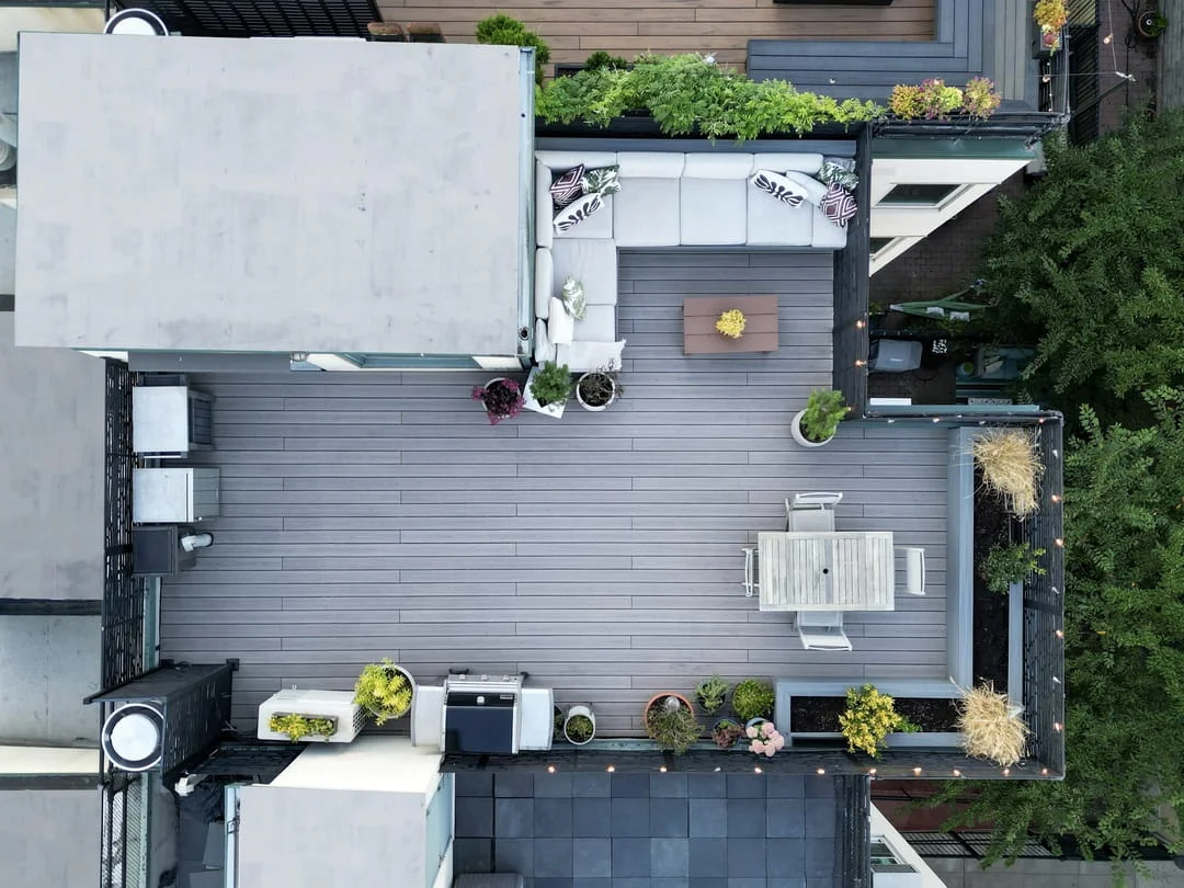 Aerial drone view of a completed Brooklyn rooftop deck with composite decking, sectional lounge seating, dining table, grill station, planters, and string lights
