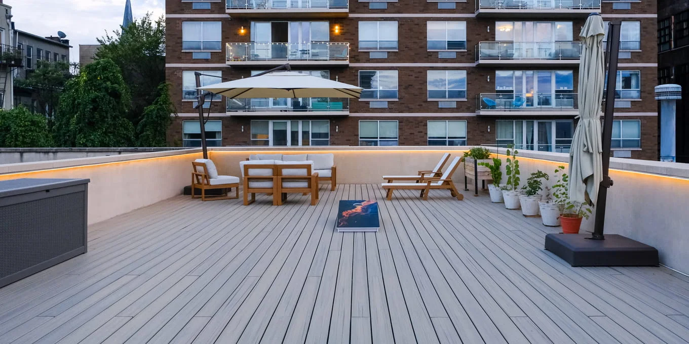Light gray composite rooftop deck with teak furniture, cantilever umbrella, and LED-lit parapet at dusk in NYC
