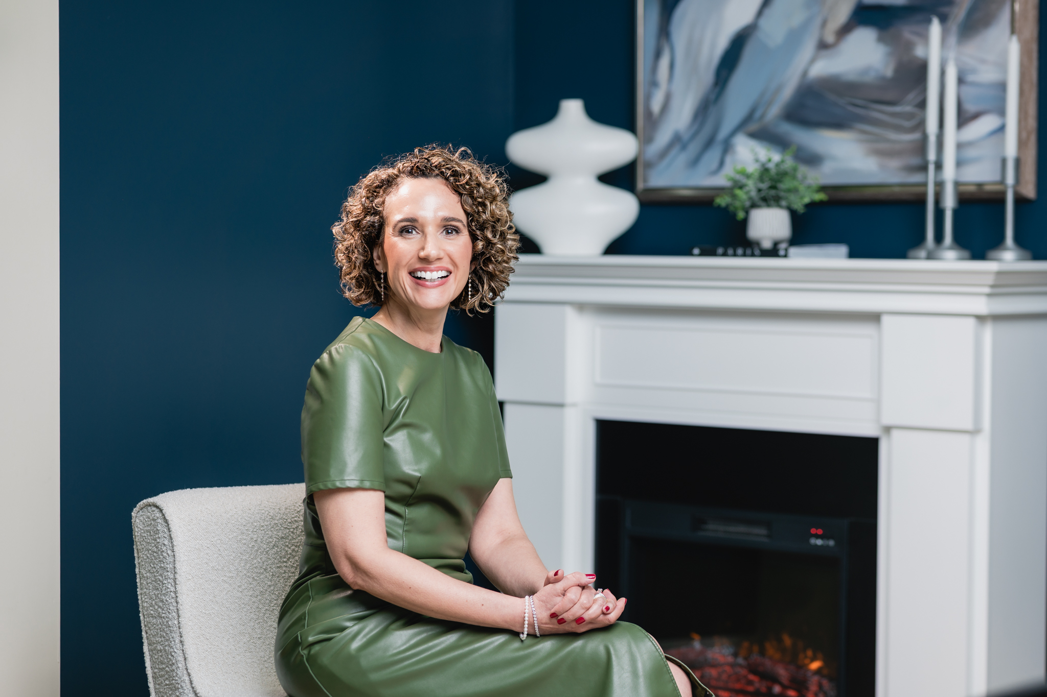 Smiling woman with curly hair wearing a green dress sitting on a chair in a room with a white fireplace and dark blue wall.