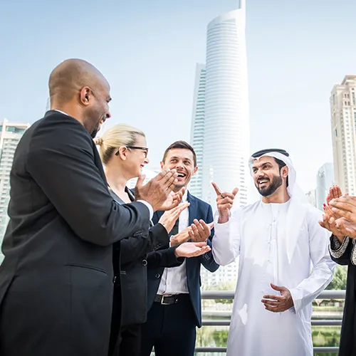 A group of multinational business professionals, including an Arab man in traditional attire, celebrating outdoors. They are exchanging handshakes and smiles, suggesting the completion of a successful deal.