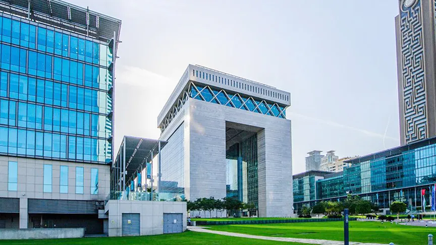 Two individuals sitting at a desk with their hands clasped, reviewing financial charts displayed on multiple monitors. The graphs on the screens show fluctuating stock or market data, with red and green lines indicating rises and falls in the market.