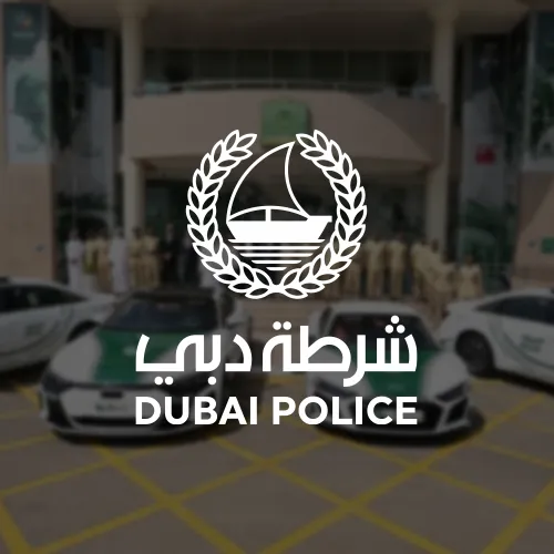 The Dubai Police emblem, depicting a sailboat encircled by laurel branches, placed in front of Dubai Police patrol cars parked outside a station.
