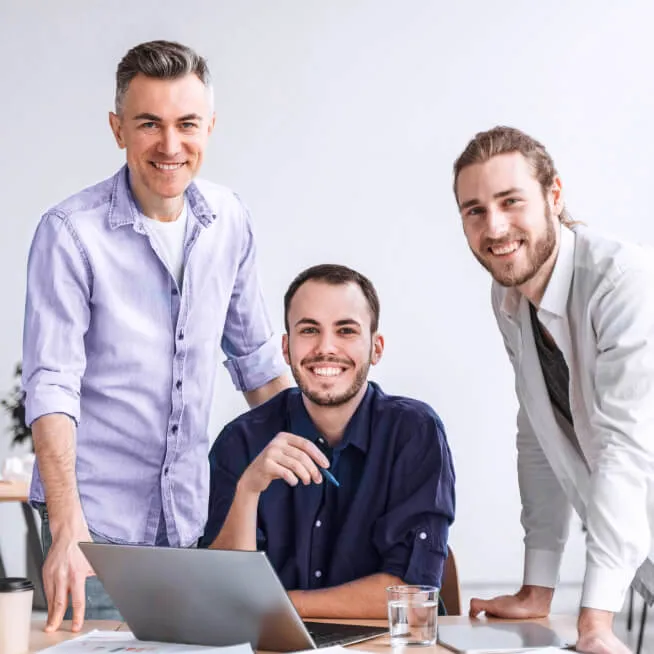 Three smiling colleagues standing around a laptop, collaborating in a bright office space, representing teamwork and a positive work environment.